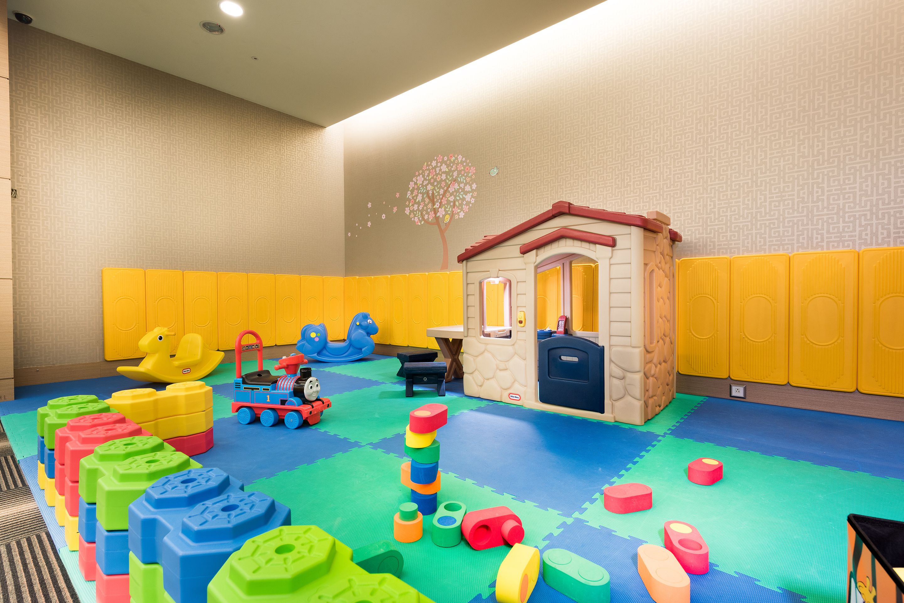 Un espace de jeux avec des blocs de mousse colorés, un train jouet et une petite maison de jeu.