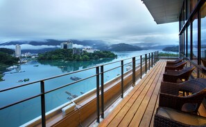 Une terrasse en bois, meublée de meubles en osier, surplombe une rivière paisible où naviguent des bateaux, le tout étant entouré de bâtiments et de montagnes.