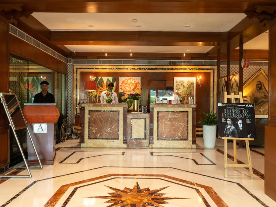 Zona de recepción de hotel con piso de mármol, un mostrador de recepción, una silla de ruedas y un cartel publicitario de un espectáculo.