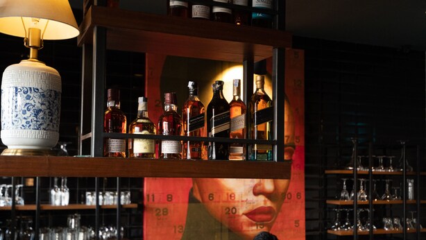 Un espace bar avec des étagères remplies de bouteilles d’alcool, un barman préparant des boissons et une lampe décorative.