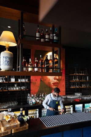 Un espace bar avec des étagères remplies de bouteilles d’alcool, un barman préparant des boissons et une lampe décorative.