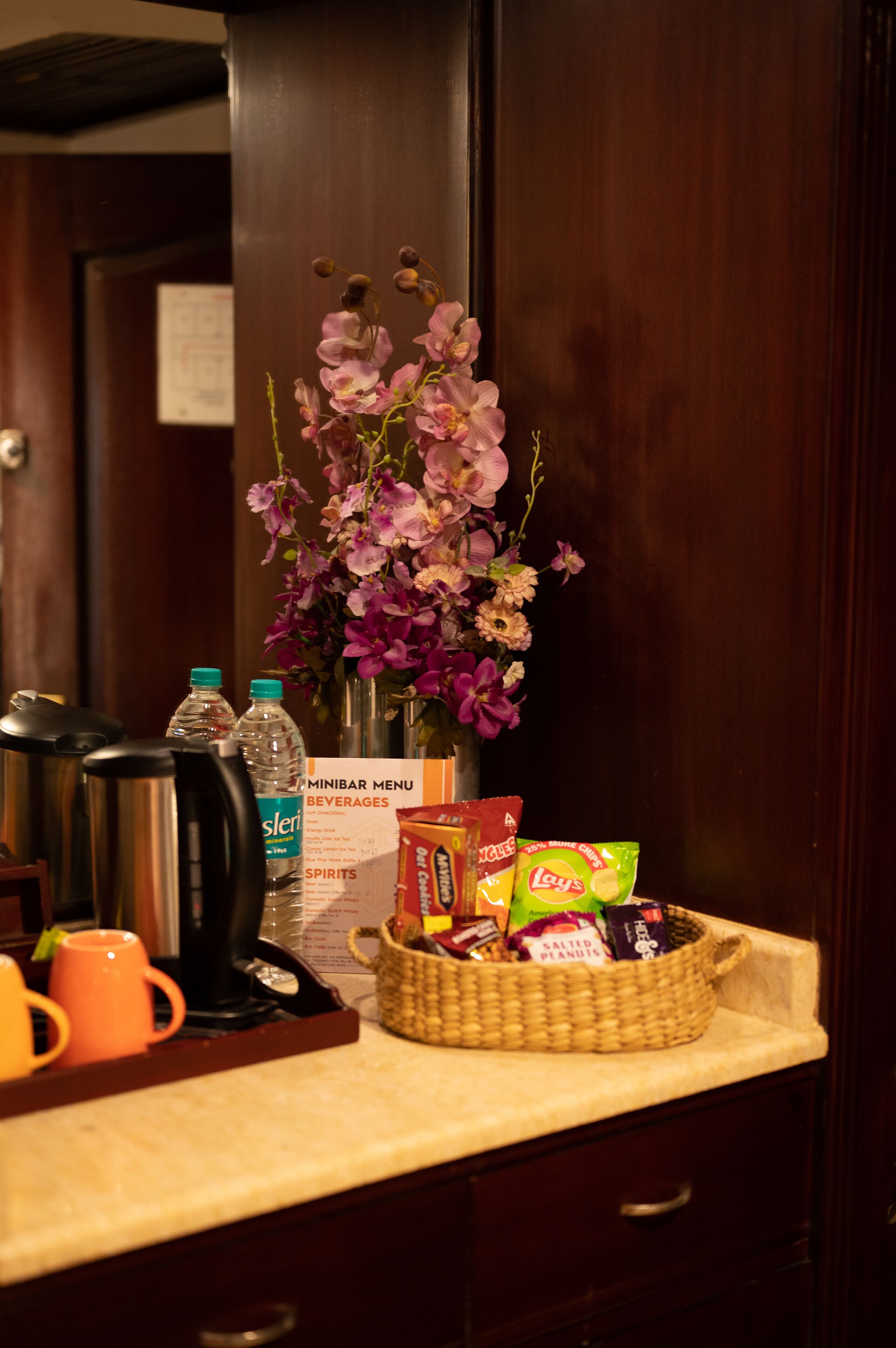 Un minibár de hotel con una variedad de bebidas, aperitivos y un jarrón decorativo con flores.