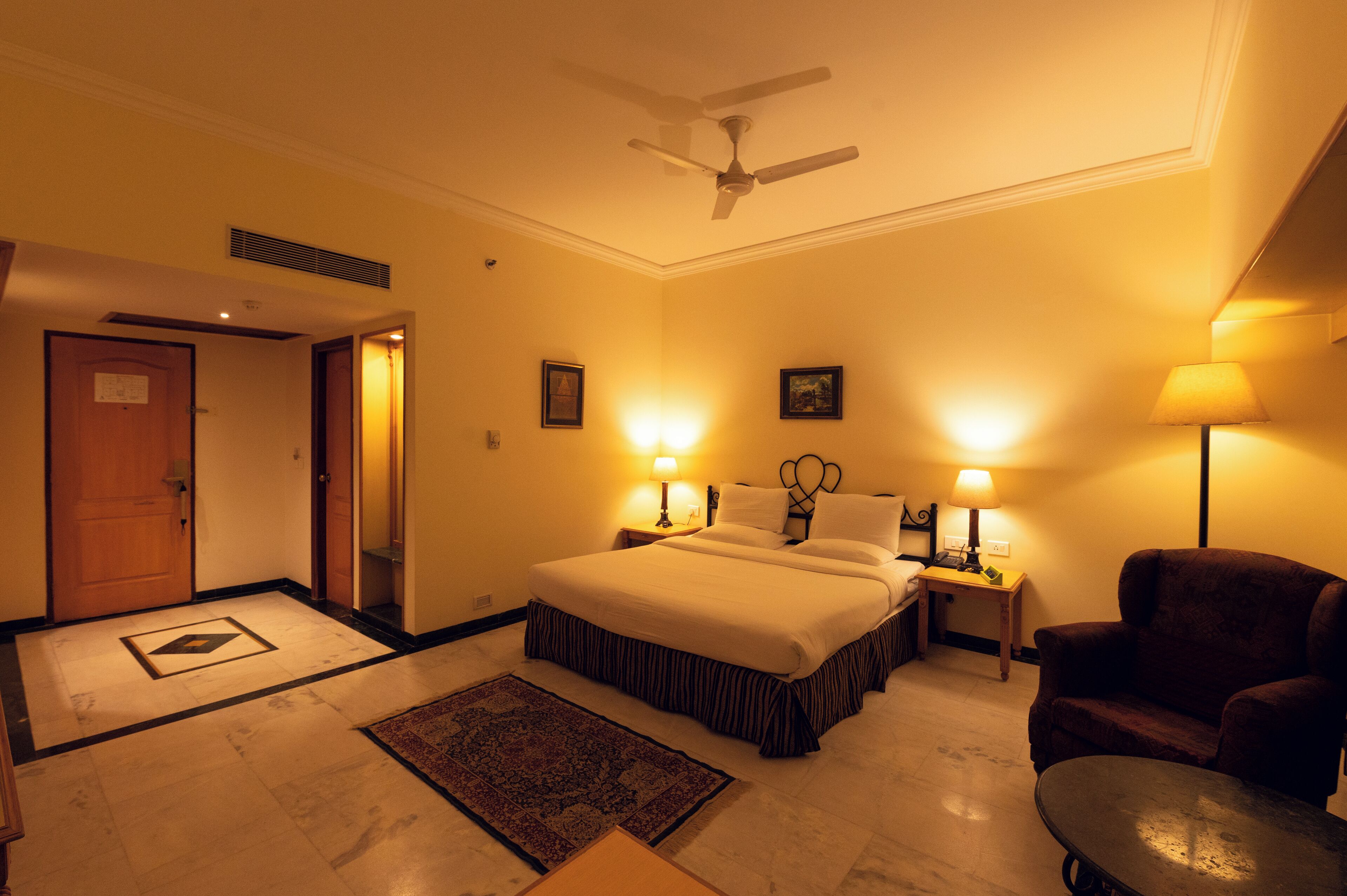 A hotel room with a bed, two bedside tables with lamps, a chair, a ceiling fan, and a door.