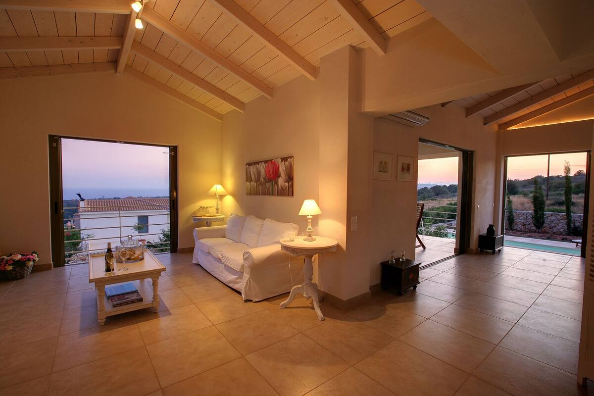 A spacious living room with a white sofa, a small table with a lamp, and a view of the outdoors through large windows.