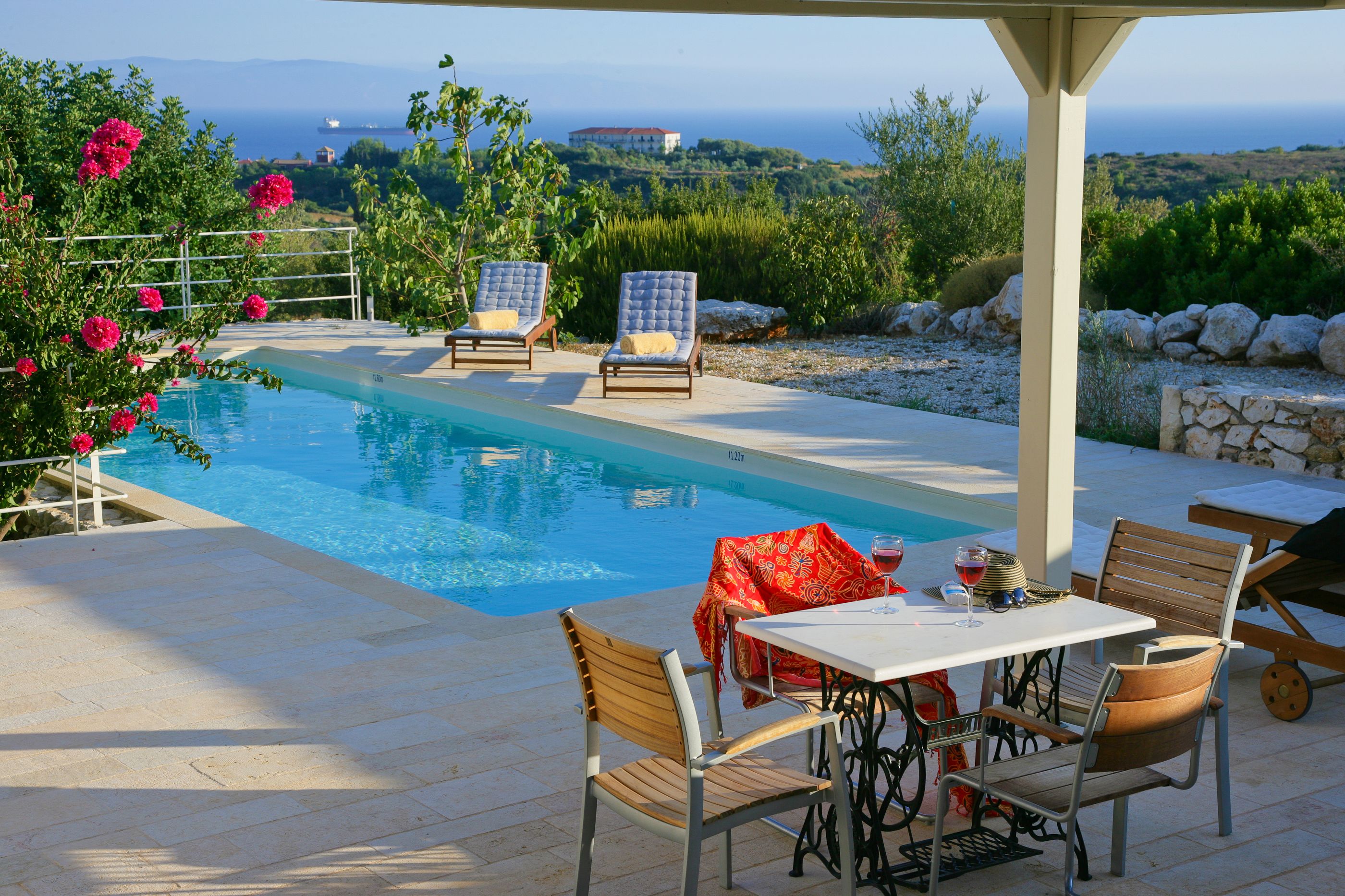 A poolside area with a table and chairs, a covered seating area, and a view of the sea.