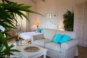 Quarto bem iluminado com cama, sofá branco, mesa de cabeceira e planta em vaso.
