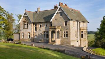 Stately manor with a central entrance, flanked by two wings, set amidst a well-maintained lawn and gravel driveway.