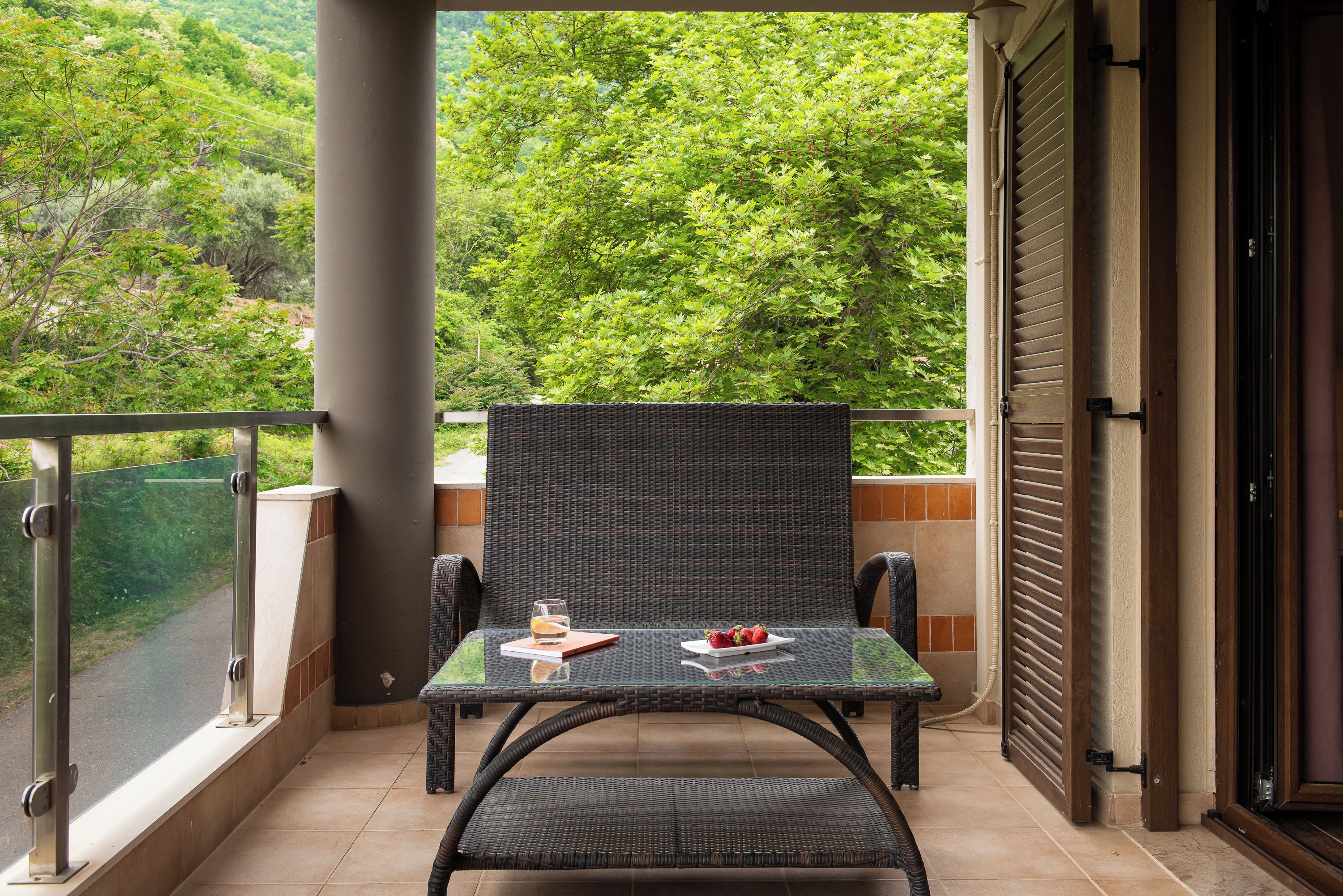 A balcony with a wicker chair and table, a glass door, and a view of lush greenery.