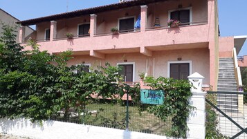 A pink building with multiple windows and balconies, surrounded by greenery and a white fence.