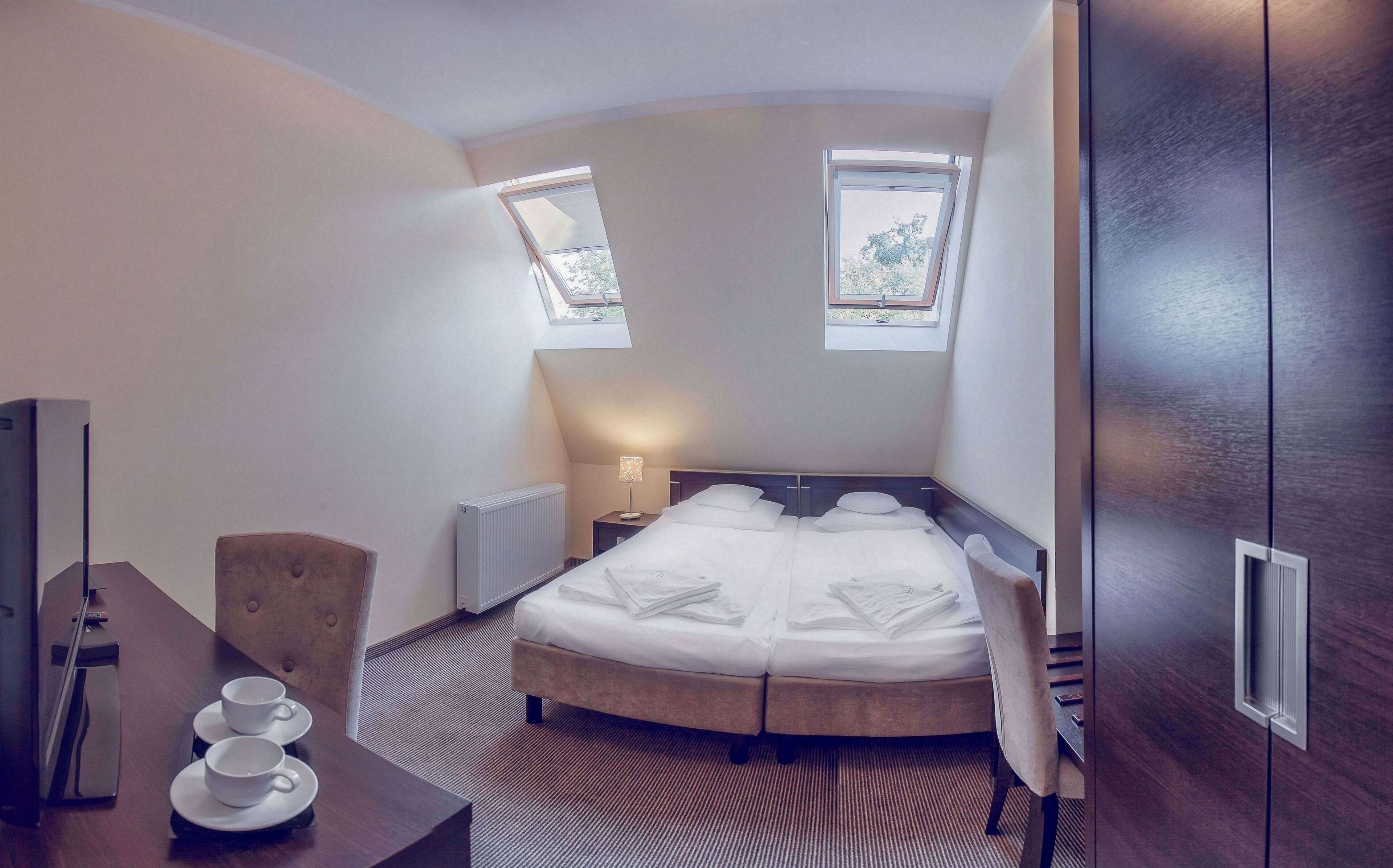 A hotel room with a large bed, two windows, a desk with a chair, and a radiator.