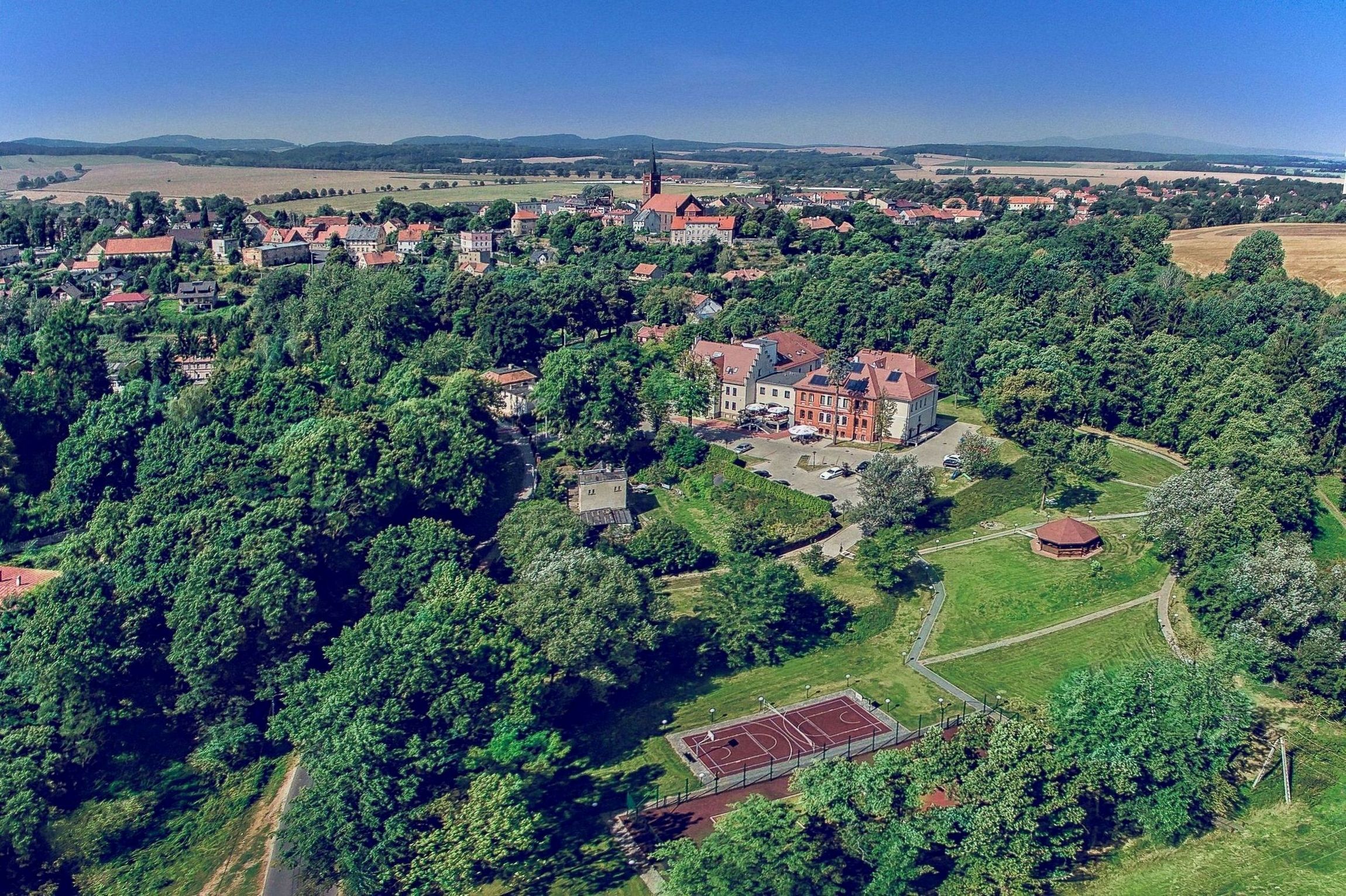Vista aerea di una città con un edificio imponente simile a un castello, circondato da vegetazione lussureggiante e un campo sportivo.