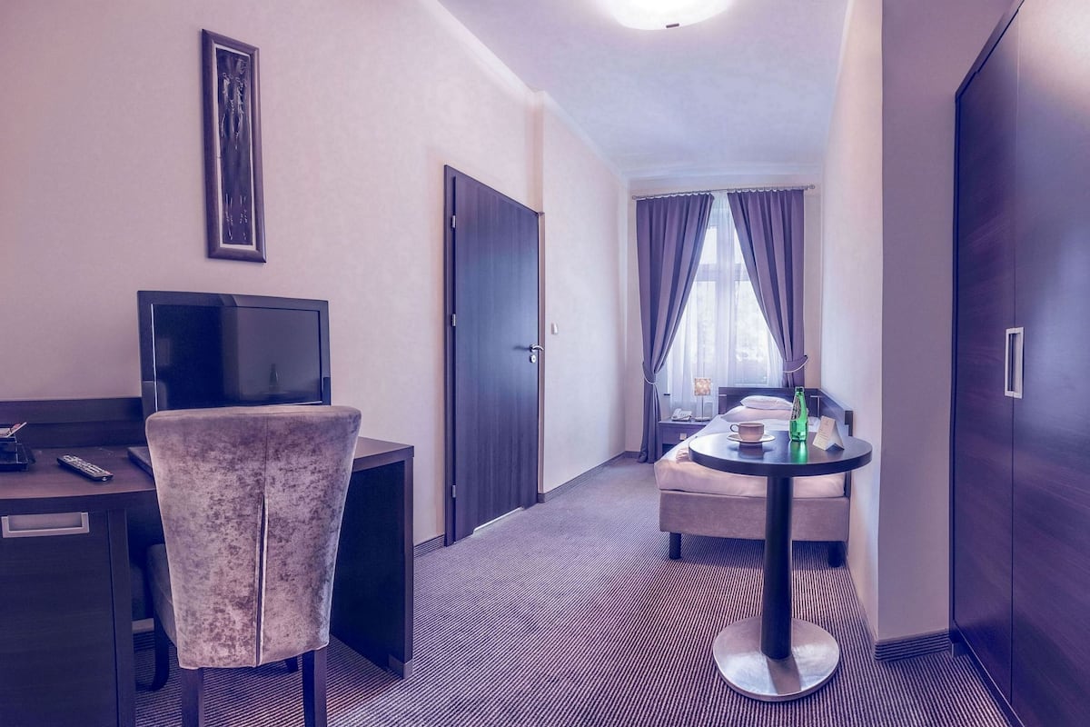 Habitación de hotel con cama, escritorio con televisor, una silla, una mesita con una botella y un armario.