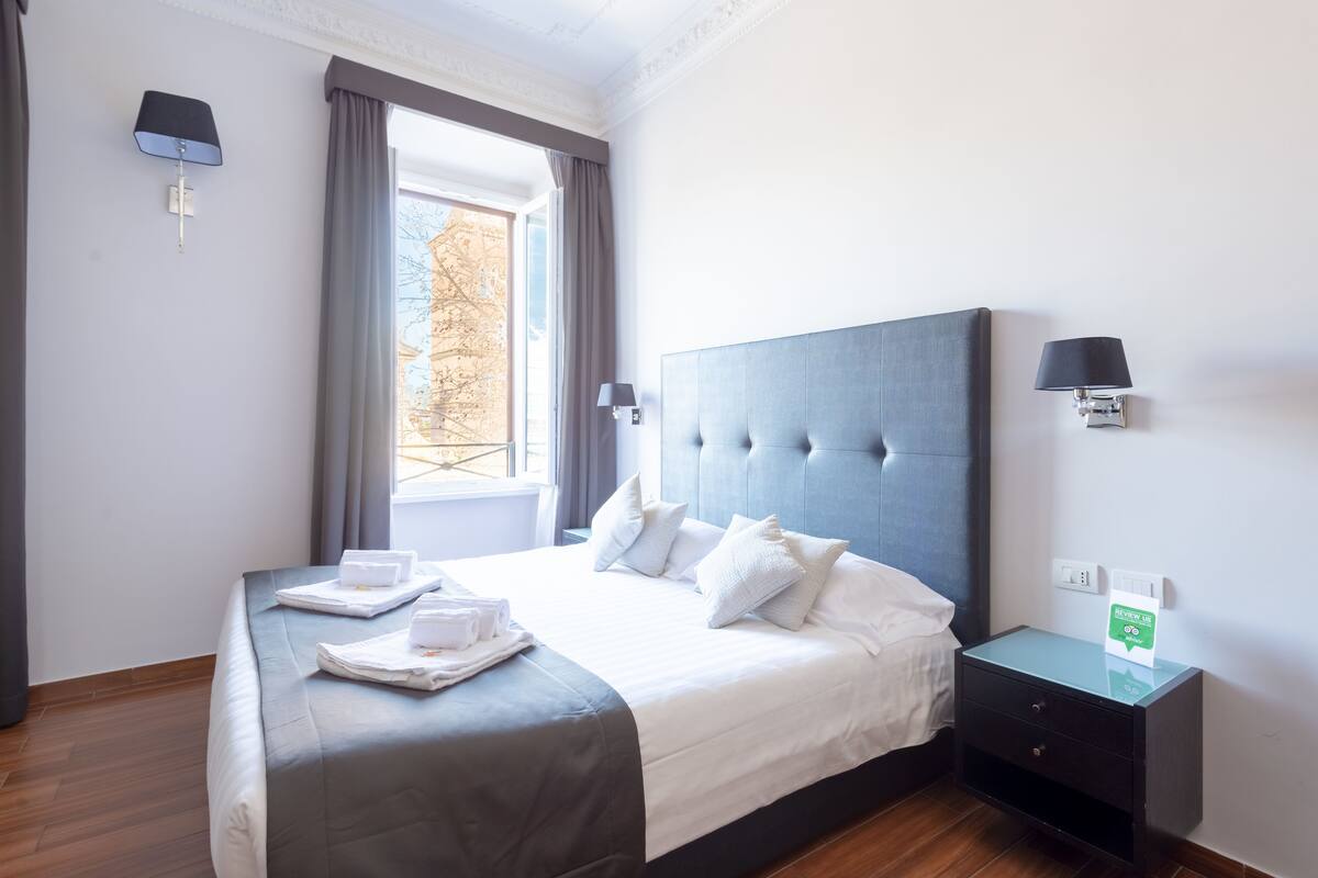 A neatly made bed with white linens and pillows, a bedside table with a lamp, and a window with curtains.