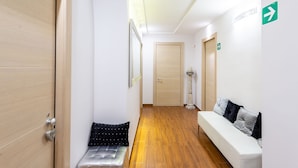 Un pasillo con suelo de madera, un banco, un sofá blanco con almohadas y una puerta con letrero de salida.