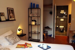 A hotel room with a bed, a nightstand with a lamp, a bookshelf, and a bathroom visible through an open door.