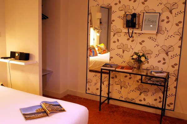 A hotel room with a bed, a bedside table with a book and a vase of flowers, a mirror, and a wall with floral wallpaper. - Decoh Studio Dragon (Marseille)