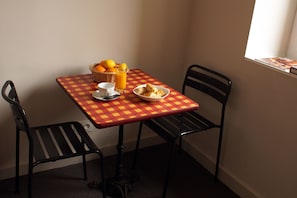 A table with a checkered tablecloth, a basket of oranges, a glass of orange juice, a plate with pastries, and two chairs.