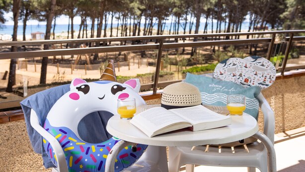 A beachside table with a book, a glass of orange juice, and a beach-themed pillow. - RVApartaments La Pineda (Torroella de Montgrí)