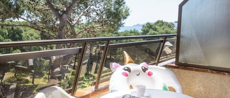 Un balcón con una silla blanca, una mesa con un libro y vasos, y un cojín con forma de unicornio.