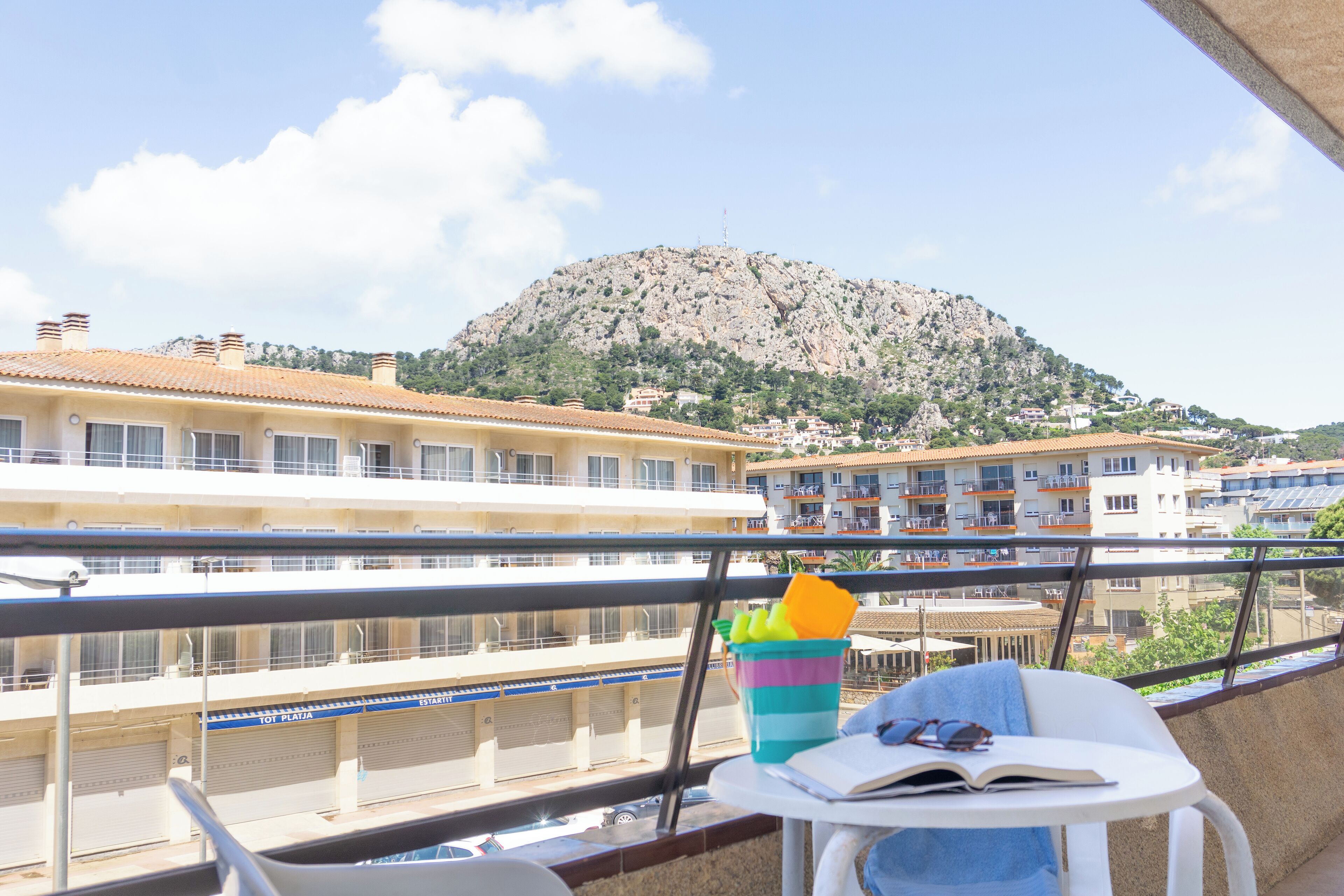 A balcony with a view of a mountain, a chair, a table with a book and sunglasses, and a colorful cup.