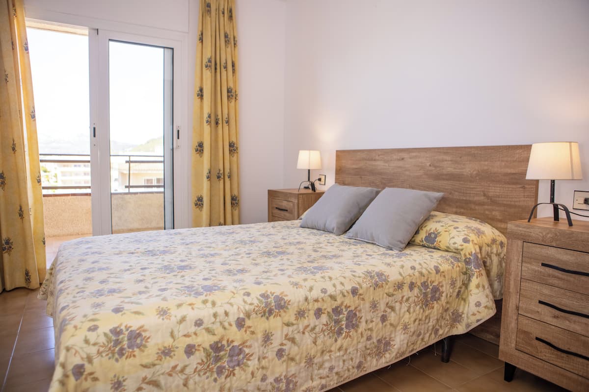 A bedroom with a bed, two bedside tables with lamps, a wooden headboard, and a sliding glass door leading to a balcony.