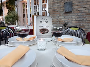 Une table dressée pour deux personnes, avec des verres à vin, des assiettes et des serviettes. On peut apercevoir, au fond, une enseigne indiquant « B.B.Q House Teraca Pasticeria ».