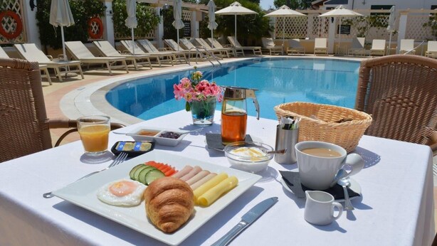 Ontbijttafel met croissants, eieren en fruit, een kan sinaasappelsap en een kop koffie bij een zwembad.