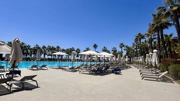 A poolside area with lounge chairs and umbrellas, palm trees, and a clear blue sky.