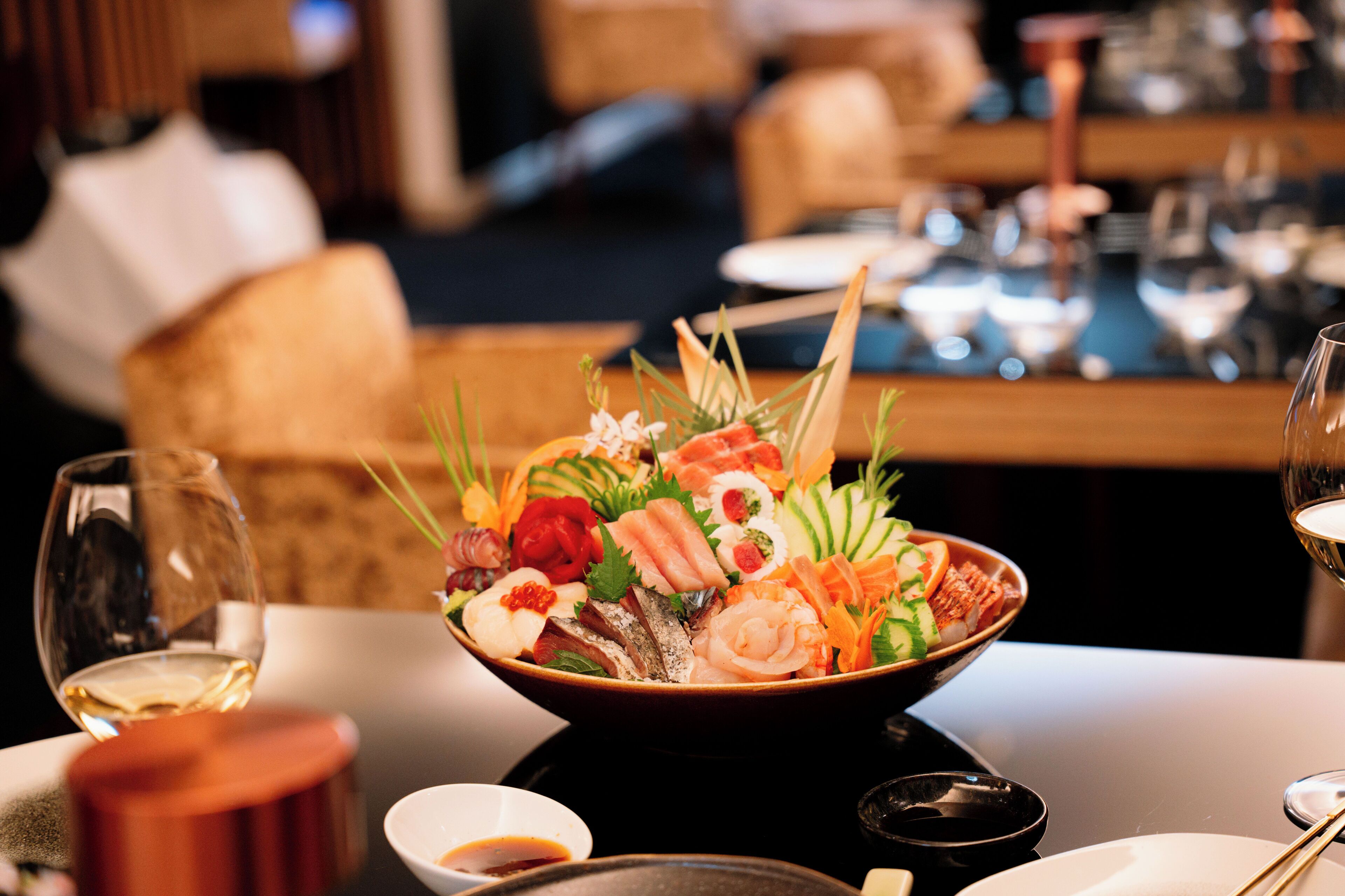 A bowl of assorted seafood and vegetables on a table with a wine glass and chopsticks.