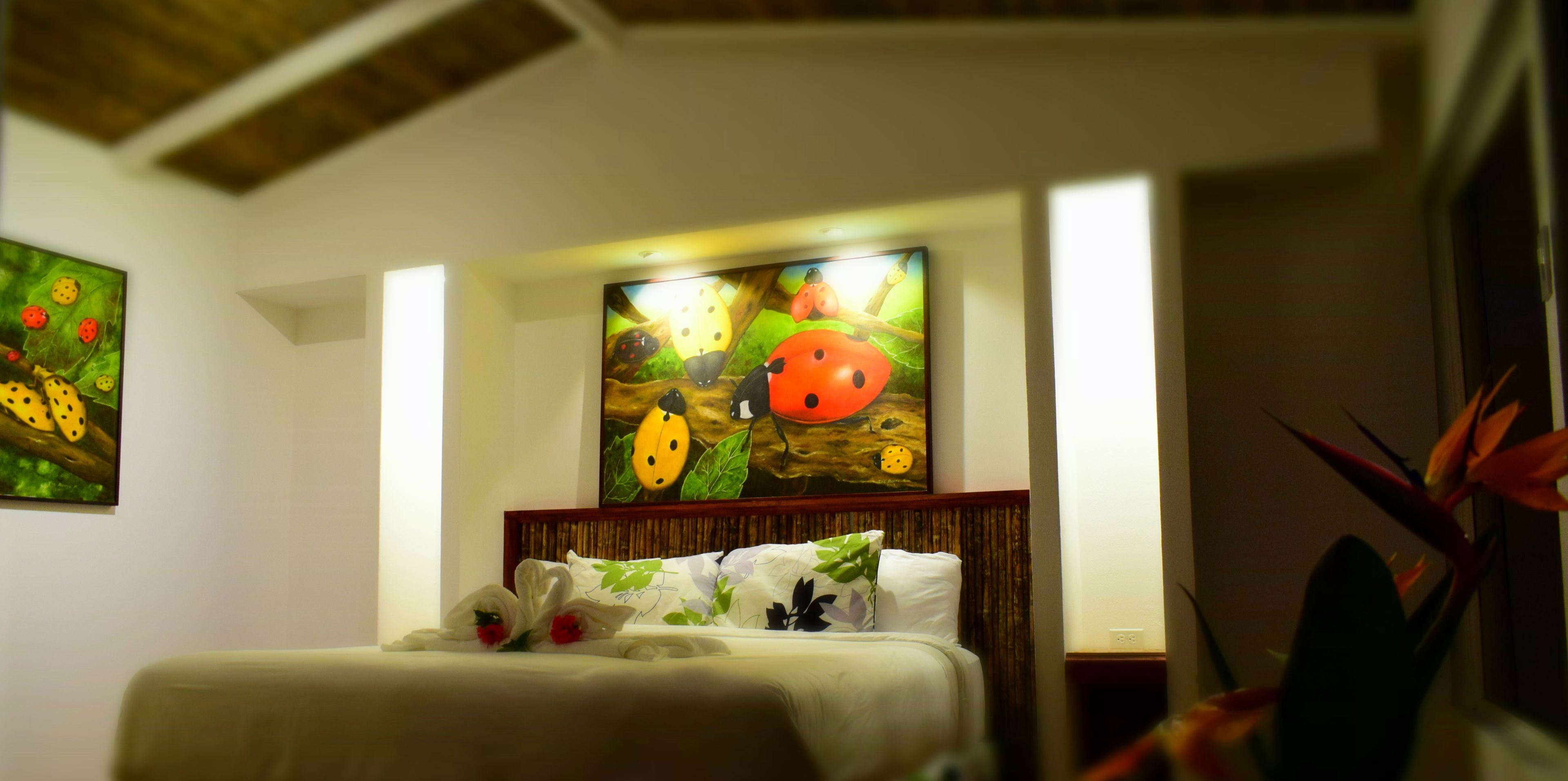 A hotel room with a bed, a painting of ladybugs on a tree branch, and a plant in the corner.