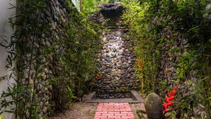 A narrow brick pathway leading to a stone wall with a small waterfall, surrounded by dense bamboo and tropical plants. - Hotel Quelitales (Cachi)