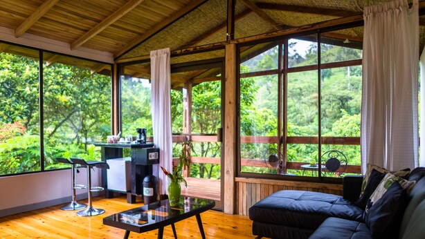 A room with a wooden ceiling, large windows, a sofa, a table, and a bar area with stools. - Hotel Quelitales (Cachi)