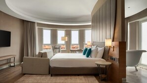 A modern hotel room with a curved ceiling, a large bed, a sitting area with a small table, a bathtub, and large windows offering a city view. - Ramada Encore by Wyndham Konya Karatay (Konya)