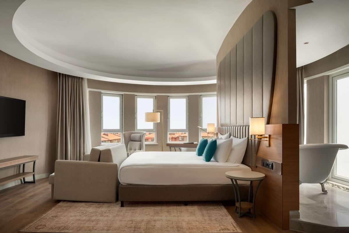 Habitació d'hotel moderna amb un sostre corbat, un llit gran, zona d'estar amb taula petita, banyera i grans finestres amb vista a la ciutat.