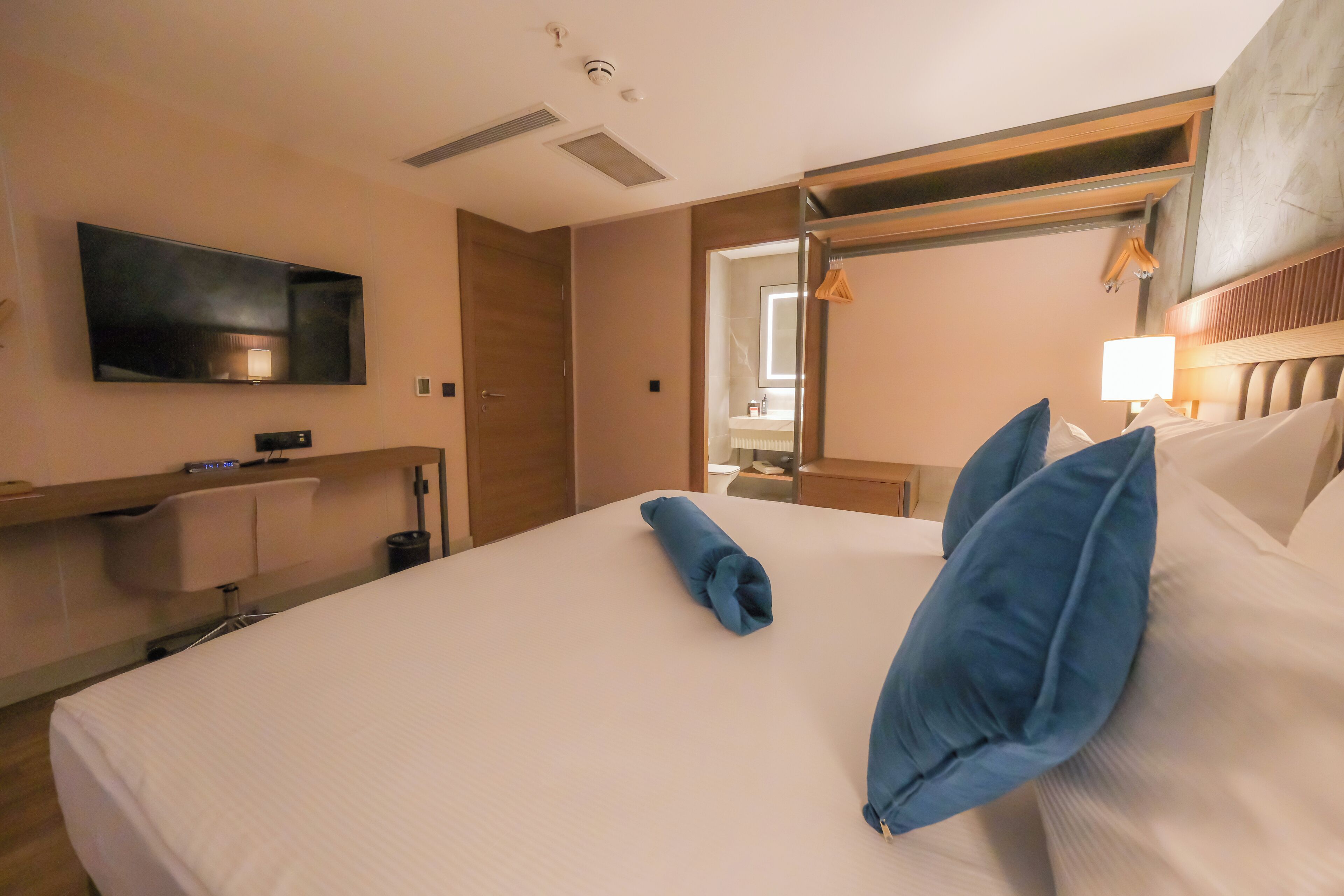 A hotel room with a large bed, a flat-screen TV mounted on the wall, a wooden headboard, and a small desk with a lamp.