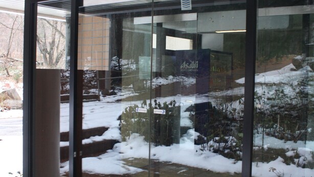 Une entrée avec une porte en verre donnant sur un espace extérieur enneigé, un bâtiment portant une enseigne « Asahi Soft Drinks », et un sol carrelé.