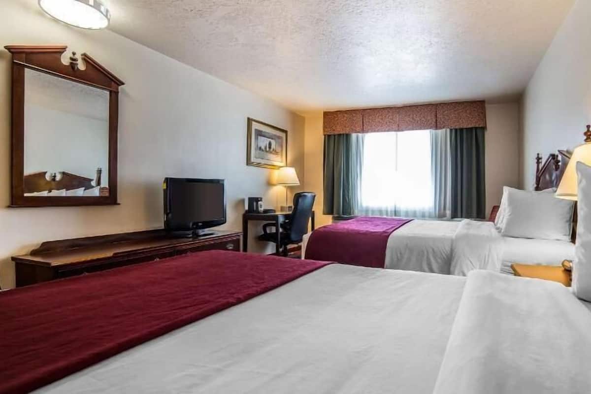 A hotel room with two beds, a desk with a television, a chair, a desk lamp, a window with curtains, and a mirror on the wall.