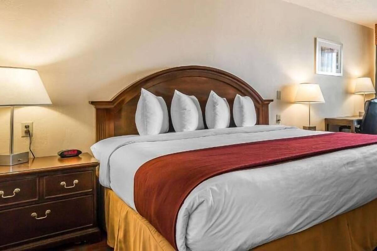 A hotel room with a bed, bedside lamps, a desk, and an air conditioning unit.