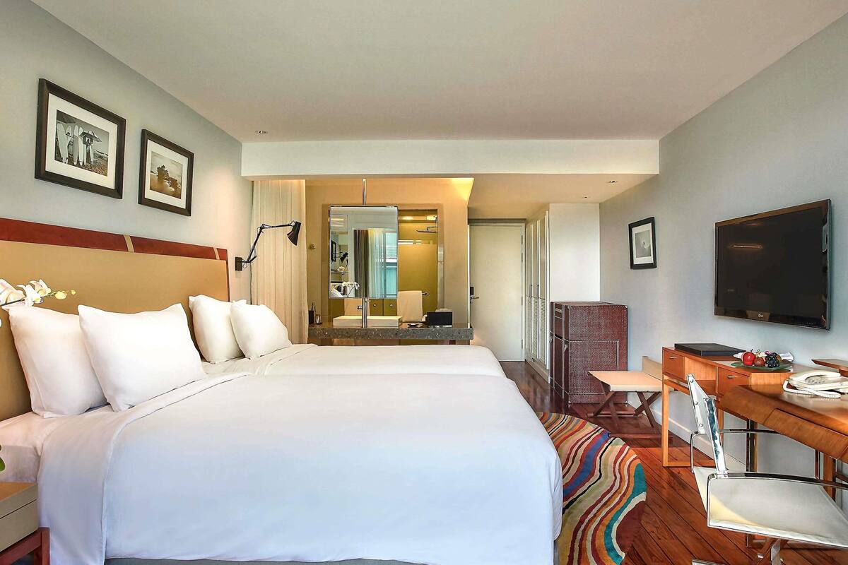 Quarto de hotel com uma cama grande, uma escrivaninha com cadeira, televisão e banheiro visível por uma porta aberta.