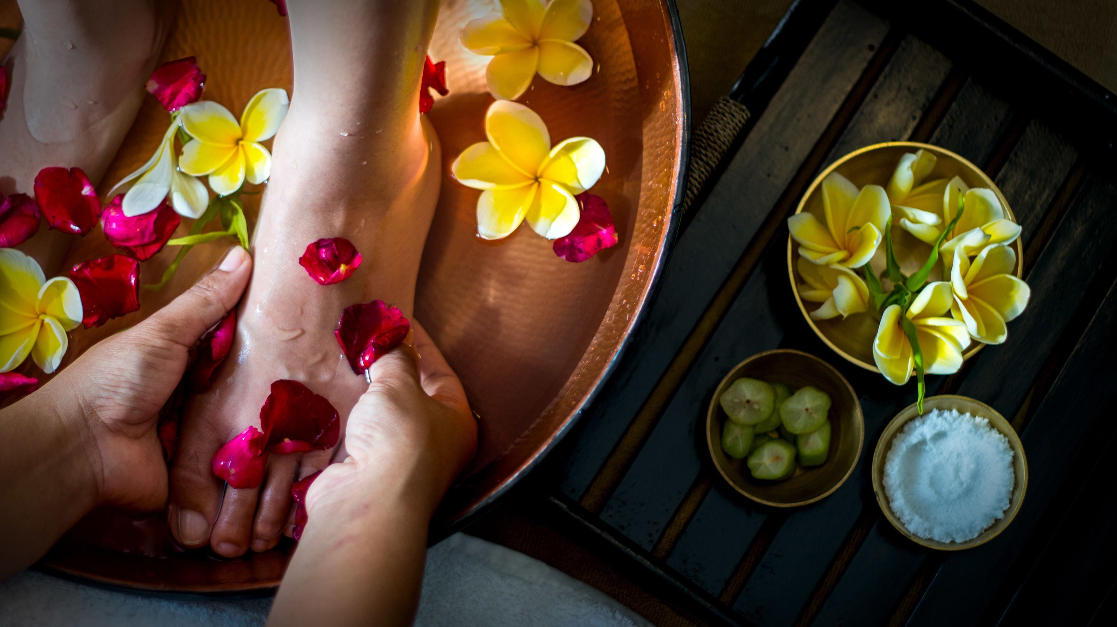 Ein hölzernes Becken mit Wasser und Blütenblättern, eine Schale mit geschnittenem Obst und eine Schale mit weißem Pulver, vermutlich für eine Spa-Behandlung.