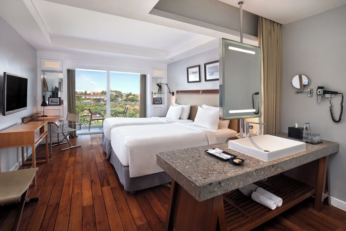 Une chambre d’hôtel avec deux lits, un bureau avec une chaise, une télévision, une grande fenêtre avec vue, une salle de bain avec un lavabo et un miroir, et un luminaire au plafond.