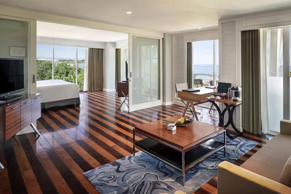 Quarto de hotel com cama, televisão, secretária com computador e vista para o oceano.