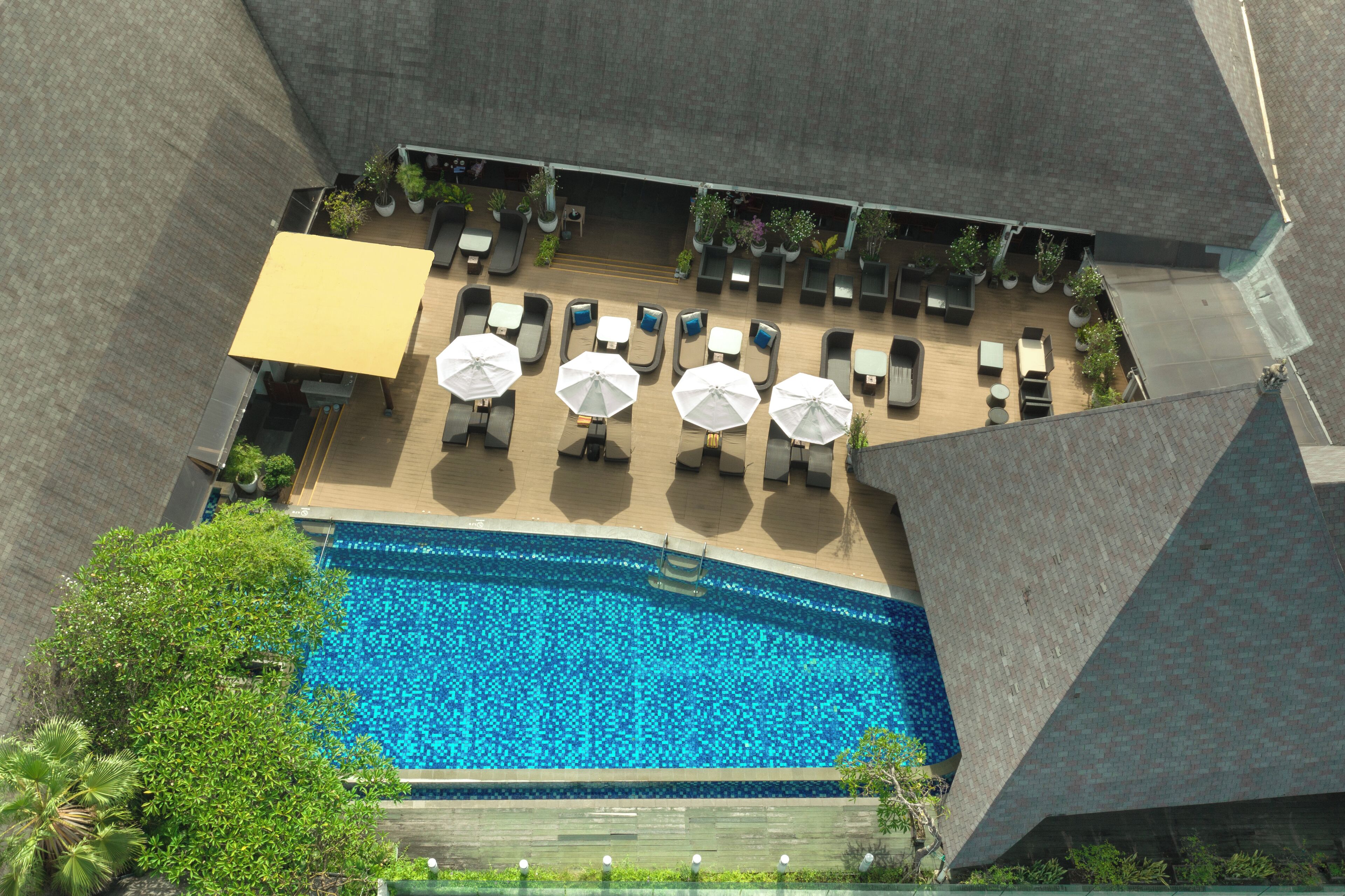 Vista aérea de una zona de piscina hotelera con tumbonas y sombrillas, rodeada de vegetación y arquitectura moderna.