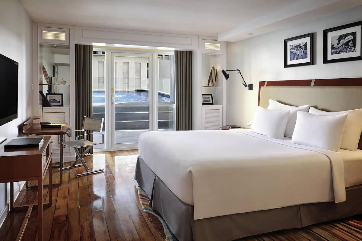 Quarto de hotel com uma cama grande, uma mesa com cadeira, televisão e porta de correr para uma varanda com piscina.