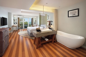 Habitación de hotel con una bañera grande, un tocador de madera, un televisor de pantalla plana y un balcón con vista al mar.