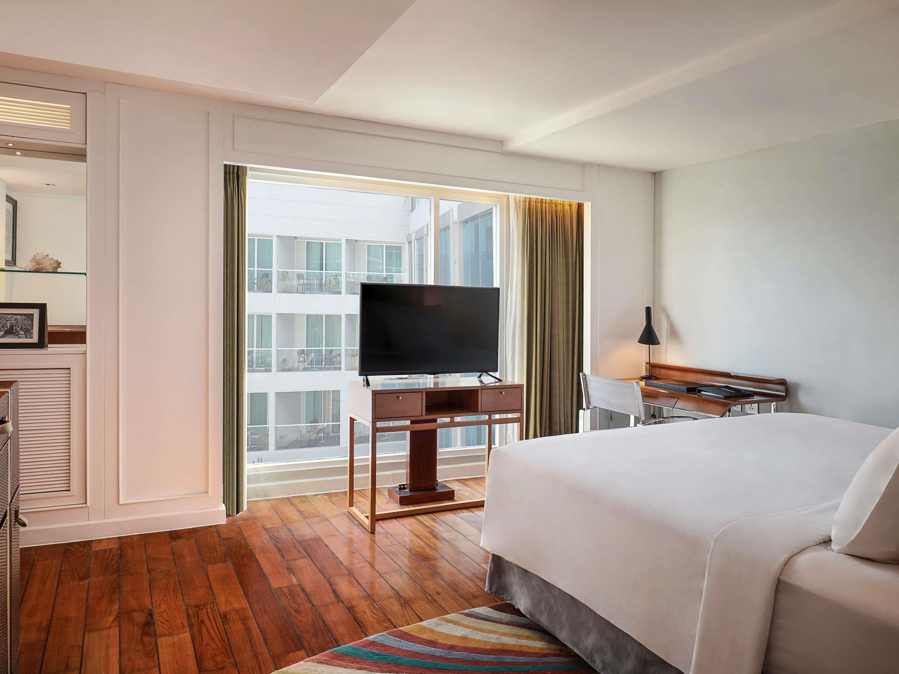 A hotel room with a large bed, a TV on a desk, a window with curtains, and a view of the building outside.