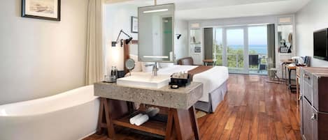 Habitación de hotel moderna con una bañera grande, un lavamanos central, un espejo amplio, una cama, un escritorio y un televisor. La habitación tiene suelo de madera y un balcón con vistas al océano.