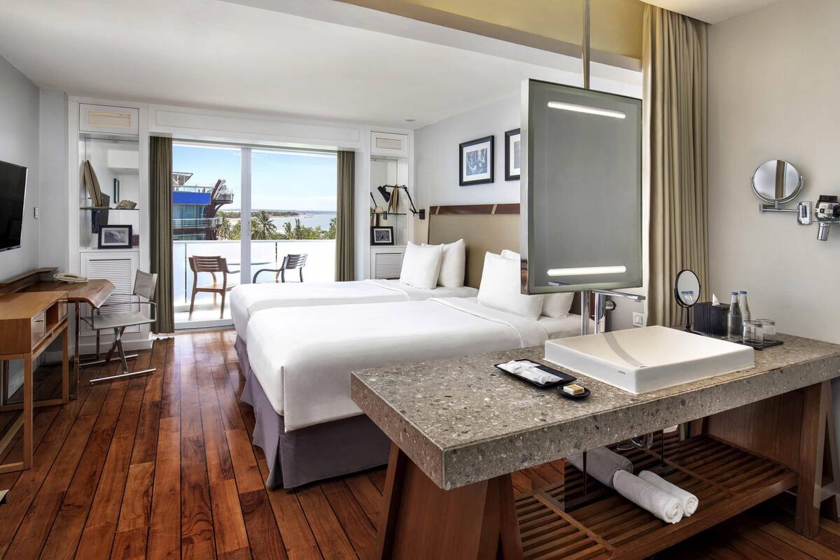 Quarto de hotel com uma cama grande, uma escrivaninha, banheiro com pia e varanda com vista.