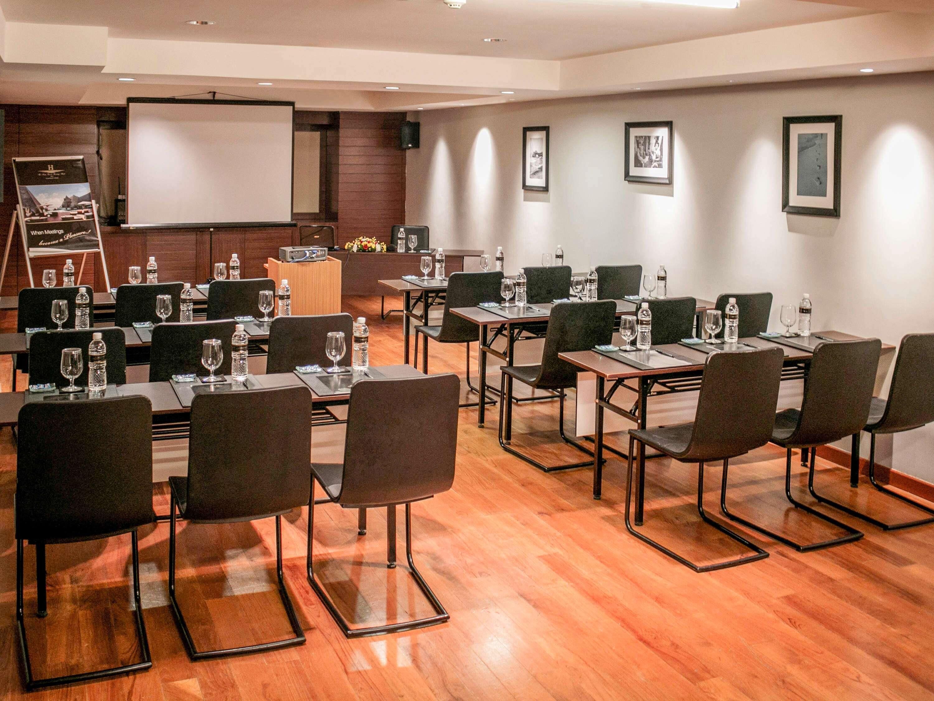 A conference room with tables, chairs, a projector screen, and framed pictures on the wall.