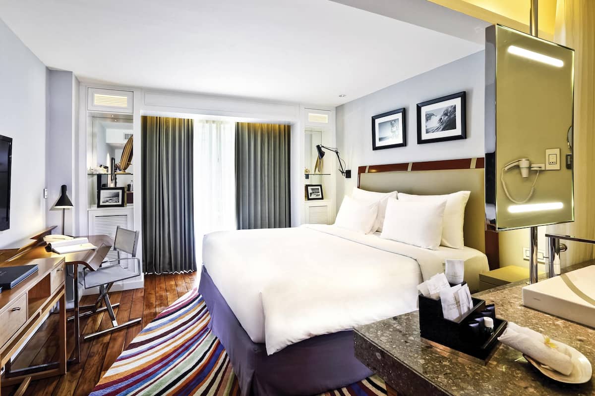 Quarto de hotel com cama, escrivaninha, cadeira e banheiro com pia e espelho.